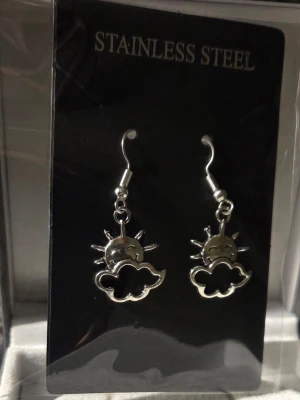 Sol och moln örhängen i stål - Snygga örhängen i rostfritt stål med motiv av en glad sol bakom ett moln. De är silverfärgade och har krokar för enkel användning. Perfekta för dig som gillar unika och lekfulla accessoarer med en touch av humor.