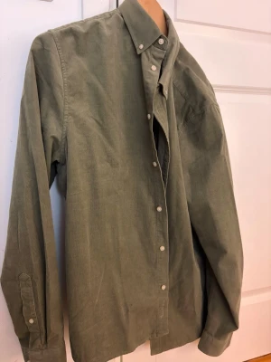 Helt Ny Grön manchester skjorta med knappar - Säljer en grön skjorta i mjuk manchester med klassisk krage och vita knappar hela vägen framtill. Skjortan har lång ärm och en avslappnad passform, perfekt för dig som gillar en stilren och enkel look. Helt ny! Ej använd. Nypris: 900kr
