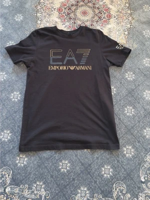 Svart EA7 Emporio Armani t-shirt - Snygg svart t-shirt från EA7 Emporio Armani med stort logotryck i guld och silver på bröstet. Klassisk rund hals och korta ärmar. Tillverkad i mjuk bomull för skön känsla och stilren look.