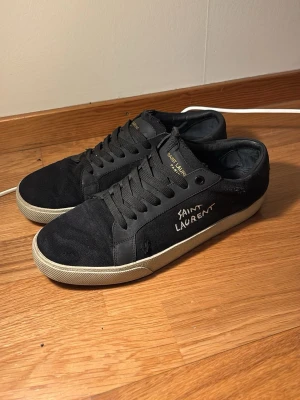 Svarta Saint Laurent sneakers - Snygga svarta sneakers från Saint Laurent med vit logga på sidan och gulddetaljer på plösen. Skorna har en klassisk låg siluett, svarta skosnören och vit sula. Materialet är en mix av canvas och läder vilket ger en stilren och exklusiv känsla.