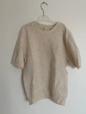 Beige stickad t-shirt med kort ärm - Mysig beige stickad t-shirt med korta ärmar och rund halsringning. Tröjan har ribbade muddar vid ärmslut och nederkant. Den är lite nopprig som man ser på bilderna men inget som märks av när man har den på sig. Lappen är tyvärr bortklippt men skulle säga att den passar en storlek S 💗