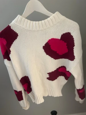 Vit stickad tröja med rosa mönster - Säljer en vit stickad tröja med stora, abstrakta mönster i mörkrött och ceriserosa. Tröjan har ribbstickad krage, ärmslut och nederkant. Oversized passform och mjukt material som känns skönt mot huden. Perfekt för dig som gillar färg och unika plagg.