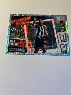 Legendary Moment fotbollskort - Säljer ett glittrigt Legendary Moment fotbollskort från Topps med Alan Shearer i Newcastle Uniteds svartvita matchställ. Kortet har holografisk yta, text och siffror för försvar och anfall samt klubbmärke och UCL-detaljer.