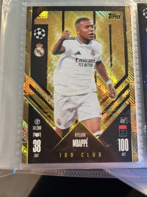 Topps fotbollssamlarbild 100 Club - Exklusiv fotbollssamlarbild från Topps med Kylian Mbappé i Real Madrid-dräkt. Kortet har en glittrig guld- och svart design med 100 Club-tryck och detaljerade stats. Perfekt för samlare av fotbollskort och sportmemorabilia.