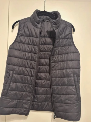 Mörkgrå quiltad dunväst - Snygg mörkgrå dunväst med quiltad design och hög krage. Västen har dragkedja framtill och två sidofickor. Tillverkad i ett lätt och smidigt material som håller värmen utan att kännas klumpig. Perfekt att slänga över en hoodie eller tröja för extra värme.