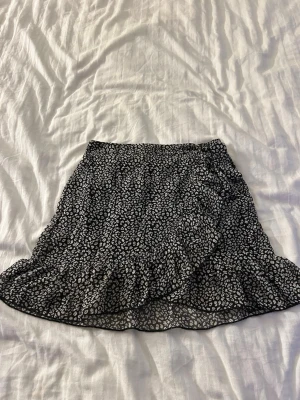 Svart omlottkjol med leopardmönster - Säljer en svart kort omlottkjol med vitt leopardmönster och volangkant. Kjolen har en luftig och flowy känsla, perfekt för dig som gillar trendiga och coola plagg. Materialet känns lätt och bekvämt, och mönstret ger en edgy vibe.