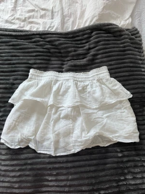 Vit volangkjol med band - Supersöt vit kortkjol med dubbla volanger och elastisk midja. Kjolen har dekorativa knytband med tofsar framtill och är gjord i ett lätt, luftigt material som passar perfekt till sommaren och våren💕Ett av banden har tappat några av pärlorna som man kan se på bilden.💕