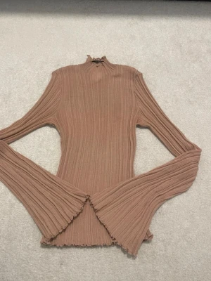 Ribbad brun stickad topp med trumpetärm - Trendig ribbad stickad topp i brun nyans med höghalsad krage och långa trumpetärmar. Materialet är mjukt och stretchigt, och kanten har en vågig finish för extra detalj. Perfekt för dig som gillar en unik och stilren look.