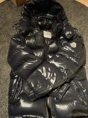 Svart dunjacka från Moncler med luva - Säljer en svart, glansig dunjacka från Moncler med stor luva och klassisk Moncler-logga på ärmen. Jackan har dragkedja framtill, två fickor med dragkedja och breda, quiltade paneler. Perfekt för kalla vinterdagar och riktigt snygg streetstil.
