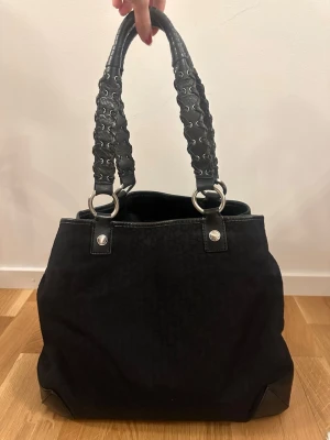 DKNY Svart handväska med flät detaljer  - Snygg svart handväska med breda handtag dekorerade med metallringar och kedjedetaljer. Väskan har en mjuk, rymlig form från DKNY. Perfekt för dig som gillar en edgy och stilren look.