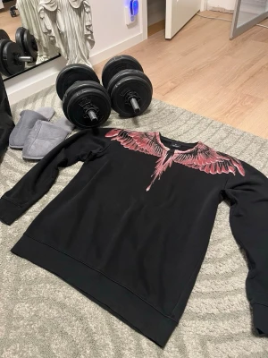 Svart sweatshirt Marcelo Burlon - Säljer en svart sweatshirt från Marcelo Burlon med ikoniskt vingmönster i rosa och rött över axlar och bröst. Tröjan har rund halsringning och är tillverkad i mjukt bomullsmaterial. Perfekt för dig som vill sticka ut med en unik design.