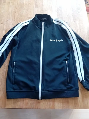 Svart Palm Angels hoodie - Snygg svart zip hoodie från Palm Angels med vita ränder längs ärmarna och logga på bröstet. Dragkedja framtill och två fickor med dragkedja. Klassisk streetwear-look med hög krage och ribbade muddar. Perfekt för dig som gillar trendiga och sportiga plagg.