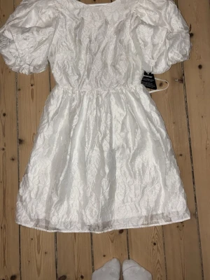 Vit kort klänning med puffärm - Superfin vit kortklänning med puffiga kortärmar och strukturmönster i tyget. Klänningen har markerad midja och rund halsringning, perfekt inför studenten! Storlek 34, ny med prislappen på🥰