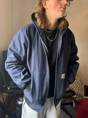 Mörkblå Carhartt hoodiejacka - Säljer en mörkblå hoodiejacka från Carhartt med dragkedja, två stora framfickor och ribbade muddar. Jackan har en klassisk Carhartt-logga på vänster ficka och är tillverkad i slitstark bomullscanvas. Perfekt för en streetwear-look.