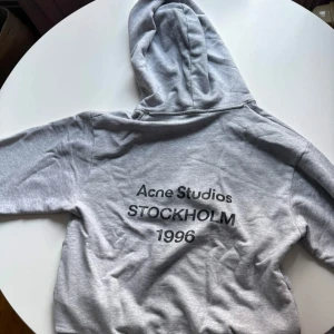 Grå hoodie från Acne Studios - Snygg grå hoodie från Acne Studios med tryck på ryggen där det står 'Acne Studios STOCKHOLM 1996'. Klassisk passform med huva och långa ärmar. Tillverkad i mjukt bomullsmaterial som känns skönt mot huden.