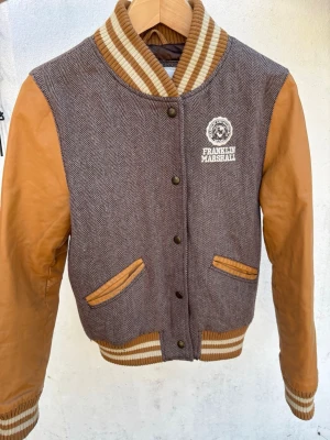 Bomberjacka Franklin & Marshall brun/beige - Snygg bomberjacka från Franklin & Marshall med brunt fiskbensmönstrat tyg och beigea ärmar i skinnimitation. Ribbstickade muddar och krage i beige och brunt. Broderad logga på bröstet och två fickor med beigea detaljer. Perfekt för dig som gillar retrostil.