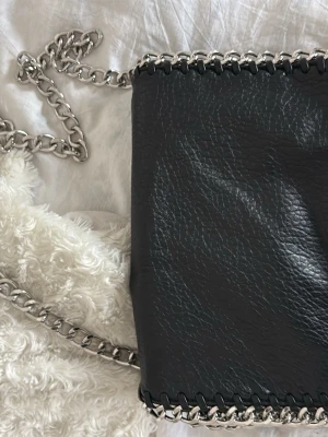 Tiamo - Svart axelväska med kedjedetaljer  - Snygg svart axelväska i fejkläder med grova silverfärgade kedjor längs kanterna och som axelrem. Väskan har en dragkedja på baksidan och en cool, lite glansig finish. Perfekt för dig som gillar en edgy och trendig look. Väskan är i den större modellen.