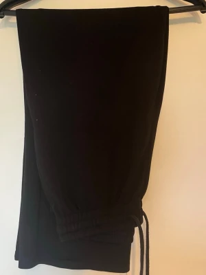 Svarta mjukisbyxor med dragsko - Ett par svarta mjukisbyxor med breda ben och elastisk midja med dragsko. Byxorna har en avslappnad passform och är tillverkade i mjukt material, perfekta för chill dagar. Klassisk och enkel design utan synliga loggor.