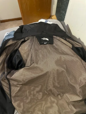 Svart vindjacka från The North Face - Snygg svart vindjacka från The North Face med huva och meshfoder på insidan. Jackan har dragkedja framtill och är tillverkad i lätt, vindtätt material. Perfekt för blåsiga dagar och enkel att packa ner i väskan.