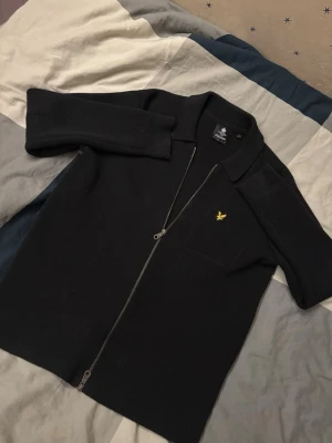 Svart stickad ziptröja Lyle & Scott - Mörk stickad ziptröja från Lyle & Scott med klassisk krage och hel dragkedja framtill. Tröjan har ett gult broderat logomärke på bröstet och är gjord i ett mjukt stickat material. Enkel och stilren design.