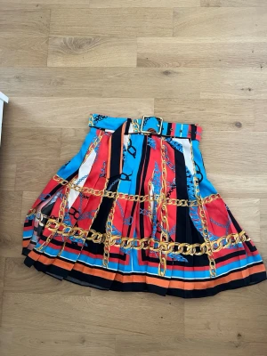 Färgglad kjol med kedjemönster Zara - Säljer en färgglad kjol från Zara i storlek XS. Kjolen har ett coolt mönster med kedjor och rep i guld, blått, rött, svart och orange. Den är veckad och har ett matchande bälte med guldfärgat spänne. 