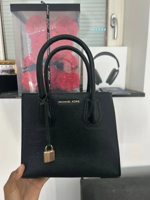 Svart handväska från Michael Kors - Snygg svart handväska från Michael Kors i strukturerat läder. Väskan har två handtag, avtagbar axelrem och guldfärgade detaljer, inklusive ett dekorativt hänglås framtill. Perfekt storlek har ej använt den. Kommer tyvär ej med mk förpakningen 