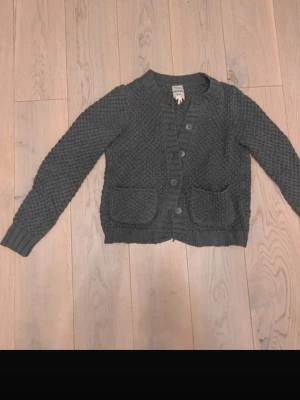 Mörkgrå stickad kofta från Monki - Mysig mörkgrå stickad kofta från Monki med grovstickad struktur. Koftan har knappar framtill, två stora fickor och ribbade muddar vid ärmslut och nederkant. Perfekt för lager på lager och en chill look.