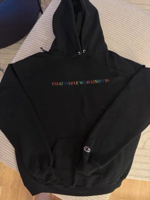 Harry Styles Treat People With Kindness Champion hoodie med brodyr - Svart hoodie från Champion med färgglad brodyrtext 'Treat People With Kindness' på bröstet. Klassisk känguruficka framtill och Champion-logga på ärmen. Mjuk och skön bomullsblandning, perfekt för en chill och avslappnad stil.