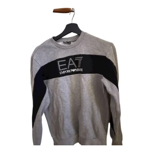 Grå sweatshirt EA7 Emporio Armani - Snygg grå sweatshirt från EA7 Emporio Armani med svart panel över bröstet och logga i vitt. Tröjan har rund hals och långa ärmar, perfekt för en chill och stilren look. Mjuk bomullskvalitet och klassisk passform. Den är stor i storleken.