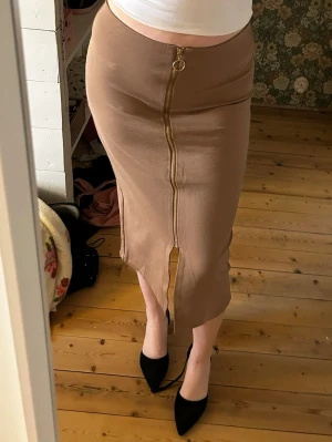 Beige midikjol med dragkedja - Trendig beige midikjol med markerad guldfärgad dragkedja framtill och slits nedtill. Kjolen har en kroppsnära passform och är gjord i ett stretchigt, mjukt material som sitter snyggt på kroppen. Perfekt för dig som gillar en stilren men edgy look.