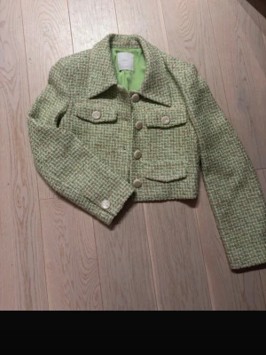 Grön rutig croppad jacka med knappar - Trendig croppad jacka i grönt och beige rutigt tyg med stora pärlemorknappar och två bröstfickor. Jackan har lång ärm och är fodrad med ljusgrönt tyg. Perfekt för dig som vill ha en chic och modern look.