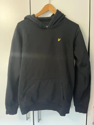 Svart hoodie från Lyle & Scott Junior - Svart hoodie från Lyle & Scott Junior med klassisk gul logga på bröstet. Hoodien har huva, känguruficka och ribbade muddar vid ärmslut och nederkant. Tillverkad i mjuk bomullsblandning, perfekt för en avslappnad och stilren look.