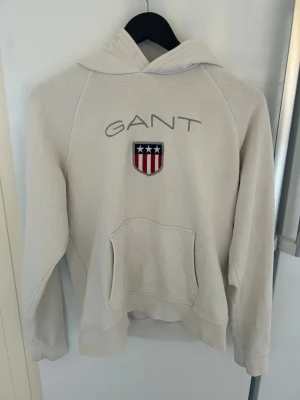 Vit hoodie från GANT med logga - Snygg vit hoodie från GANT med stor broderad logga och amerikanskinspirerat sköldmärke på bröstet. Klassisk känguruficka framtill och huva. Perfekt för dig som gillar stilrena och sportiga plagg. Mjuk bomullskänsla och avslappnad passform.