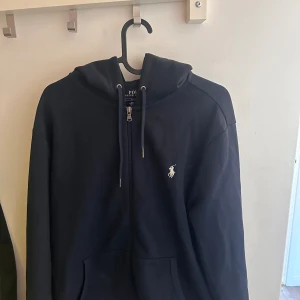 Marinblå hoodie från Polo Ralph Lauren - Snygg marinblå hoodie från Polo Ralph Lauren med dragkedja framtill och klassisk huva med dragsko. Ikoniska vita loggan broderad på bröstet. Tillverkad i mjukt bomullsmaterial och har två fickor framtill. Perfekt för en avslappnad och stilren look.