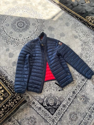 Mörkblå dunjacka från Parajumpers - Snygg mörkblå dunjacka från Parajumpers med quiltad design och röd insida. Jackan har dragkedja framtill, hög krage och en patch med logga på ärmen. Perfekt för kalla dagar och riktigt lätt att bära.