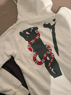 Vit Friends- hoodie med ormtryck - Säljer en vit hoodie från VLONE med stor grön FRIENDS- text på framsidan och ett fett ormtryck i rött, svart och vitt på ryggen. Hoodien har huva med dragsko och ribbade muddar. Perfekt för dig som gillar streetwear och vill sticka ut.