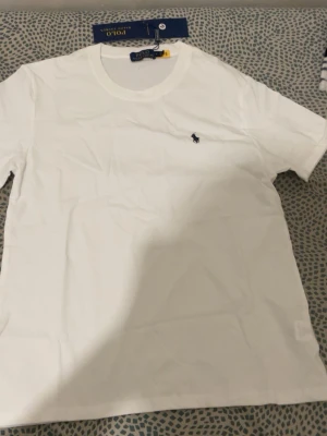 Vit t-shirt från Polo Ralph Lauren - Klassisk vit t-shirt från Polo Ralph Lauren med rund hals och den ikoniska lilla loggan broderad på bröstet. Tillverkad i mjuk bomull för en skön känsla och perfekt passform. Enkel och stilren design som funkar till allt.