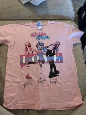 Rosa Dolly Style t-shirt med tryck - Säljer en rosa t-shirt från Dolly Style med stort färgglatt tryck av gruppen och texten LOVE på framsidan. T-shirten har korta ärmar och är i mjuk bomull. Perfekt för dig som gillar pop och vill sticka ut med en unik look.