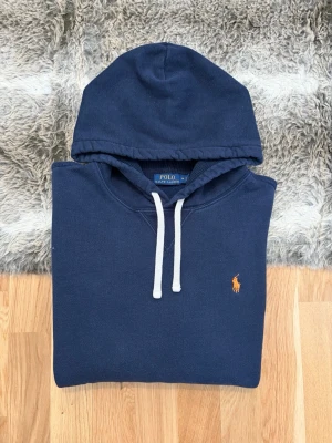 Marinblå Polo Ralph Lauren Hoodie - Storlek M - Hej! 👋 Säljer en riktigt fräsch marinblå Polo Ralph Lauren hoodie i storlek M. Den har en sjukt snygg detalj med pony-loggan broderad i kontrasterande orange på bröstet 🐎 Tröjan är i mycket gott skick, välvårdad och har den där klassiska mjuka fleece-insidan ✨ Sitter perfekt (Modell: 177cm) 👕 Modellen saknar QR-kod, men alla invändiga tvättlappar sitter kvar och intygar äktheten ✅ En tidlös favorit som funkar till allt. Fraktar snabbt! 🚛📦💬💯 