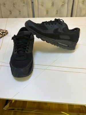 Svarta Nike Air Max 90 sneakers - Säljer ett par helsvarta Nike Air Max 90 sneakers med klassisk chunky sula och ikonisk Swoosh på sidan. Skorna är tillverkade i en mix av läder och mesh för både stil och komfort. Perfekta för dig som gillar streetwear och vill ha en tidlös look.