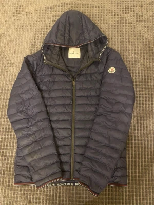 Mörkblå dunjacka från Moncler - Snygg mörkblå dunjacka från Moncler med huva och dragkedja framtill. Jackan har Moncler-logga på ärmen och diskreta detaljer i rött, vitt och blått längs huva och nederkant. Perfekt för kalla vinterdagar och riktigt lätt att bära. Storlek 2. Slitage på höger ficka, lite trög. 