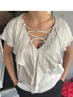 Vit topp med snörning framtill - Säljer en vit topp med korta ärmar och snygg snörning framtill i urringningen. Toppen har ett avslappnat fall och volangdetaljer längs framsidan. Materialet ser ut att vara tunt och luftigt, perfekt för varma dagar.