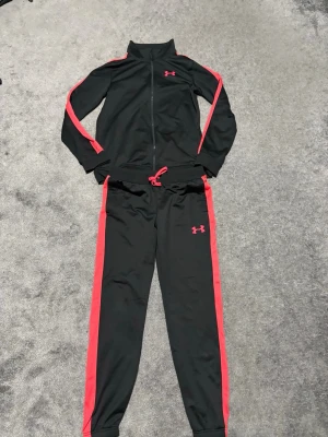 Svart träningsset från Under Armour - Säljer ett svart träningsset från Under Armour med röda detaljer längs ärmar och ben. Setet består av en zip-jacka och matchande byxor med dragsko i midjan. Perfekt för gymmet eller uppvärmning. Materialet är mjukt och stretchigt för maximal komfort.