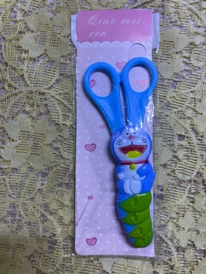 Blå barn-sax med tecknad figur - Rolig barn-sax med blå handtag och en tecknad figur på handtaget, inspirerad av en kattrobot. Figuren är blå, vit, grön och röd. Saxen är gjord av plast och metall och passar bra för pyssel och hobby.