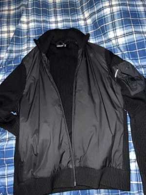 Svart cardigan J.Lindeberg  - Snygg svart cardigan från J.Lindeberg med stickade ärmar och krage. Jackan har dragkedja framtill och en ficka med dragkedja på ärmen. Materialmix av polyester och stickat ger en cool kontrast och modern look. Köpte den ny för 1800kr och är näst intill lika ny som då