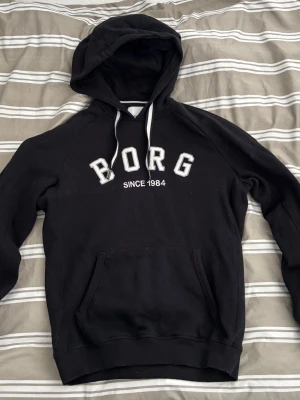Svart hoodie från Björn Borg - Svart hoodie från Björn Borg med vit text 'BORG SINCE 1984' på bröstet. Klassisk känguruficka framtill, vita dragsnören i huvan och mjukt material. Perfekt för en avslappnad och sportig stil.