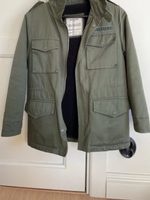 Trendig Zadig & Voltaire jacka i mycket bra kvalité!! - Snygg olivgrön parkas från Zadig & Voltaire med fyra stora fickor framtill och coola detaljer på ryggen med text av diamanter. Jackan har hög krage, dragkedja och inbyggd luva samt axelklaffar. Perfekt för dig som vill ha en praktisk men trendig jacka. Jag har endast använt den 1 gång och den är därför i väldigt bra skick!