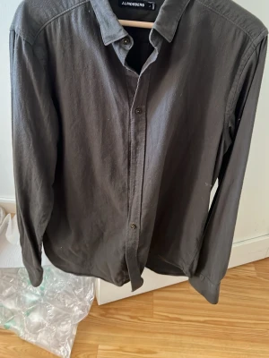 Mörkgrå skjorta från J.Lindeberg - Stilren mörkgrå skjorta från J.Lindeberg med klassisk krage och knäppning framtill. Skjortan har långa ärmar och är tillverkad i ett mjukt bomullsmaterial. Perfekt för dig som gillar en enkel men snygg look.