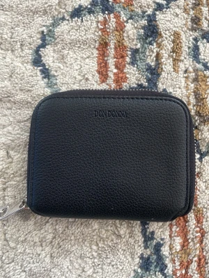 Svart plånbok från Don Donna - Snygg svart plånbok från Don Donna i ett stilrent och kompakt format. Tillverkad i strukturerat konstläder med dragkedja runtom. Perfekt för kort och kontanter, enkel att ha med sig i väskan eller fickan. Använd ett fåtal gånger. Öppen för prut, skriv gärna vid minsta fundering😁
