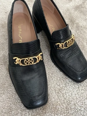 Svarta loafers med gulddetalj Unisa - Stilrena svarta loafers från Unisa i präglat lädermönster. Skorna har en fyrkantig tå, låg bred klack och en snygg guldfärgad metallkedja framtill. Perfekta för dig som gillar klassisk men trendig stil.